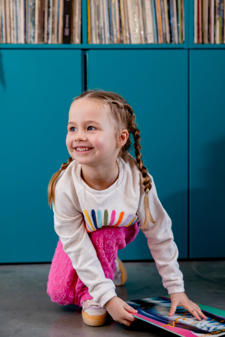 Meisje draagt ecru sweater met bloem in regenboogkleuren op een roze broek
