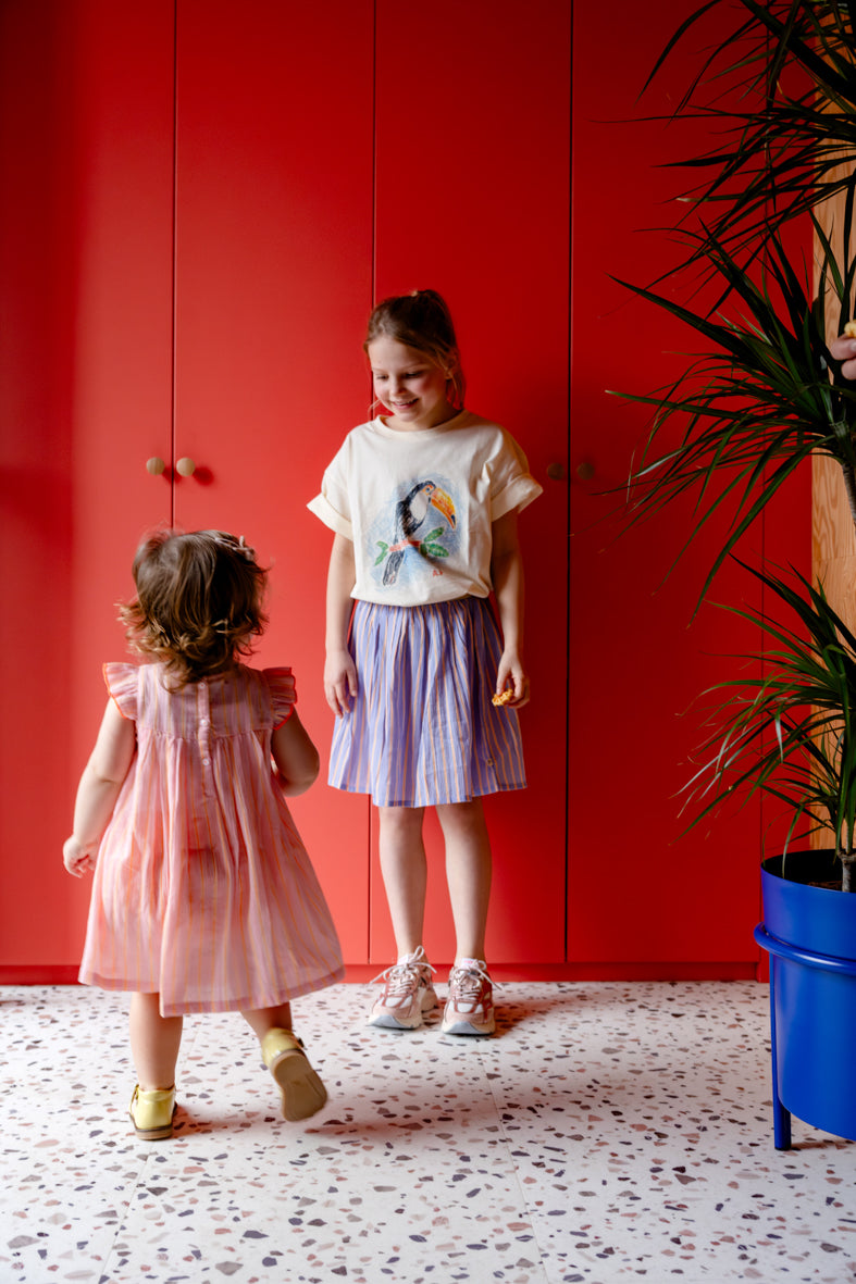 Kind draagt ecru t-shirt met toekan gecombineerd met blauwe rok met 
strepen.
