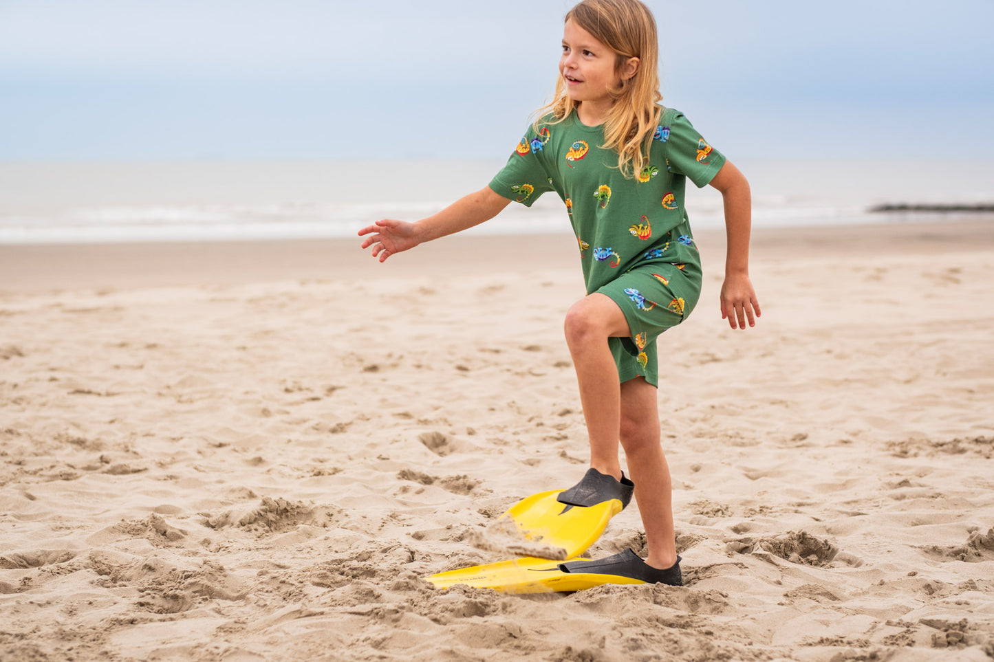 Kind draagt groene pyjama met kameleons op het strand
