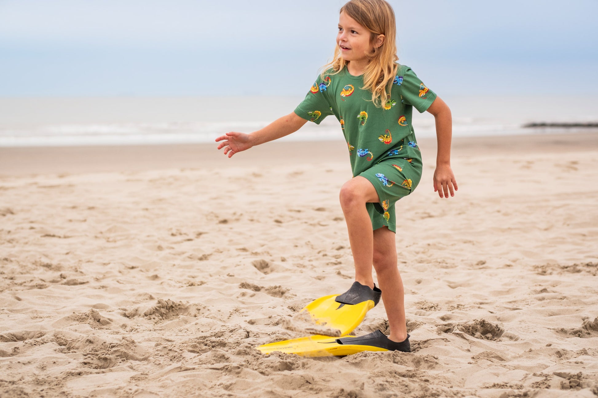 Kind draagt groene pyjama met kameleons op het strand

